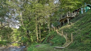 Una scala in legno conduce a una casa verde su una collina, circondata da una fitta foresta e un piccolo ruscello.