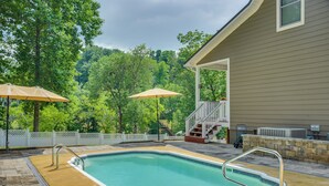 A swimming pool with a white deck, two umbrellas, and a covered area with stairs leading to a house. - Modern Tims Ford Lake Home w/ Private Dock & Pool! (Lynchburg)