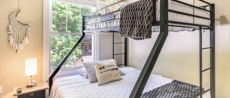A bunk bed with a white quilt, a macrame wall hanging, and a window with a view of trees.