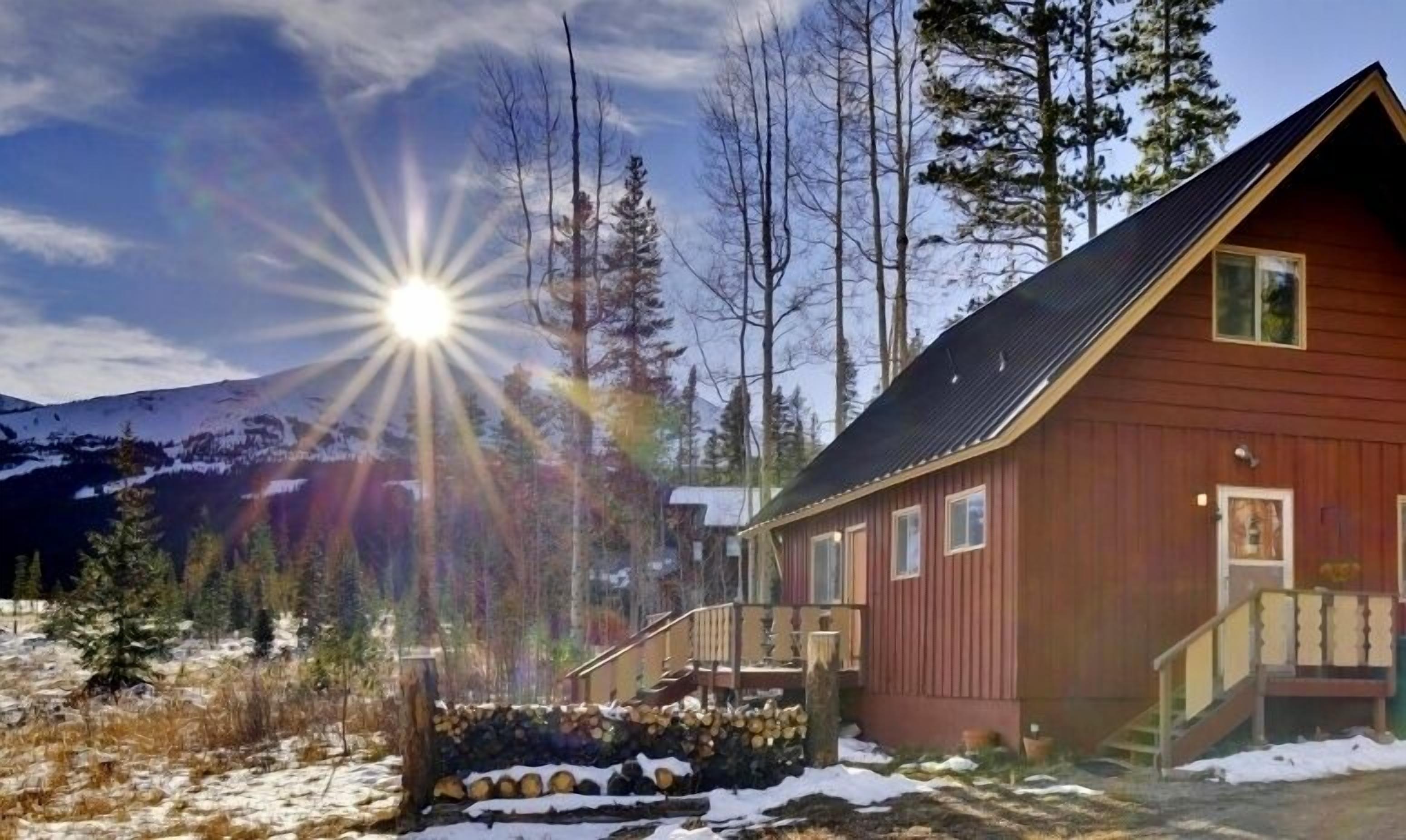 A wooden cabin with a balcony, surrounded by snow-covered ground and trees, with a mountain in the background.