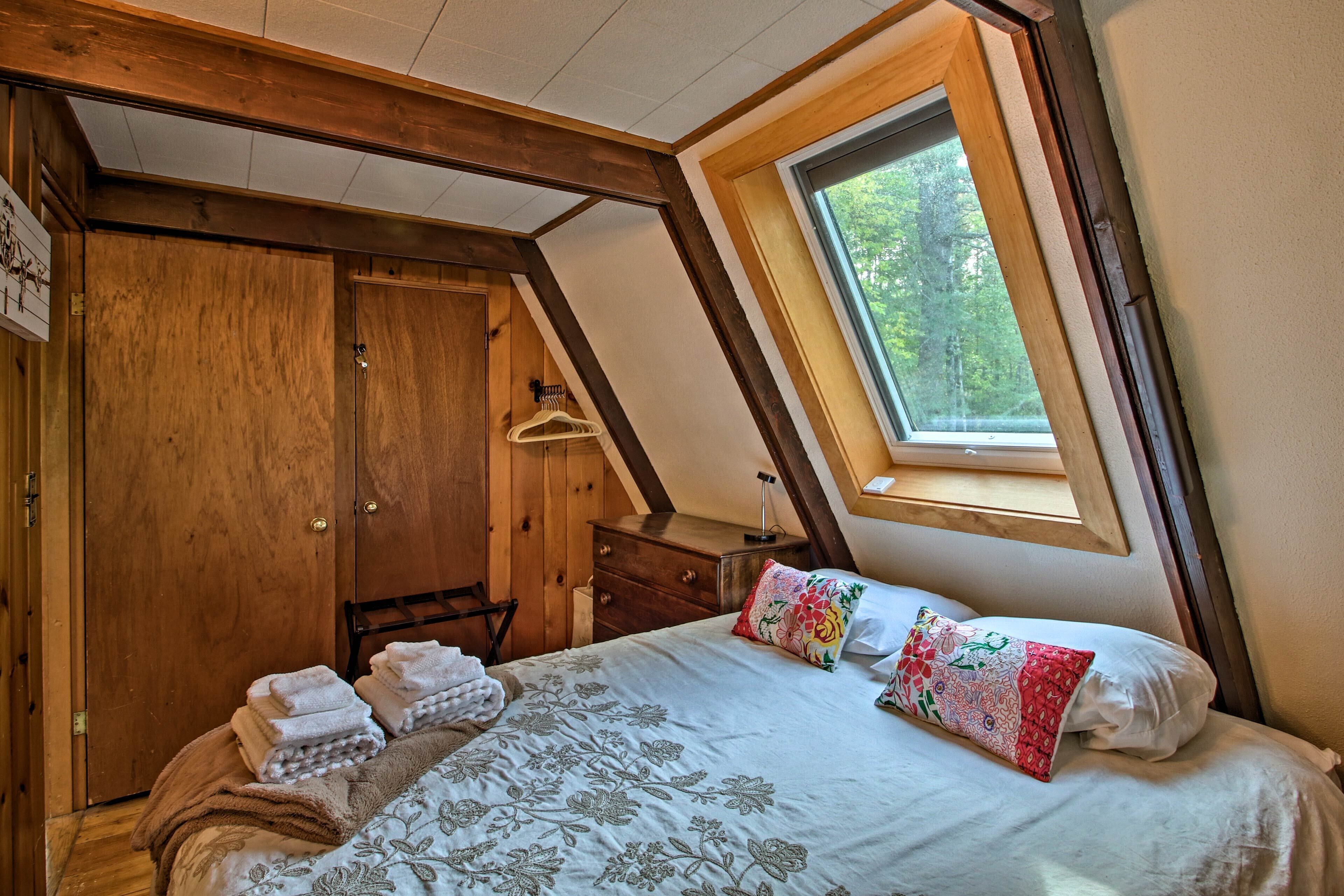 A bedroom with a sloped ceiling, wooden wardrobe, a bed with floral bedding, and a window with a view of trees.