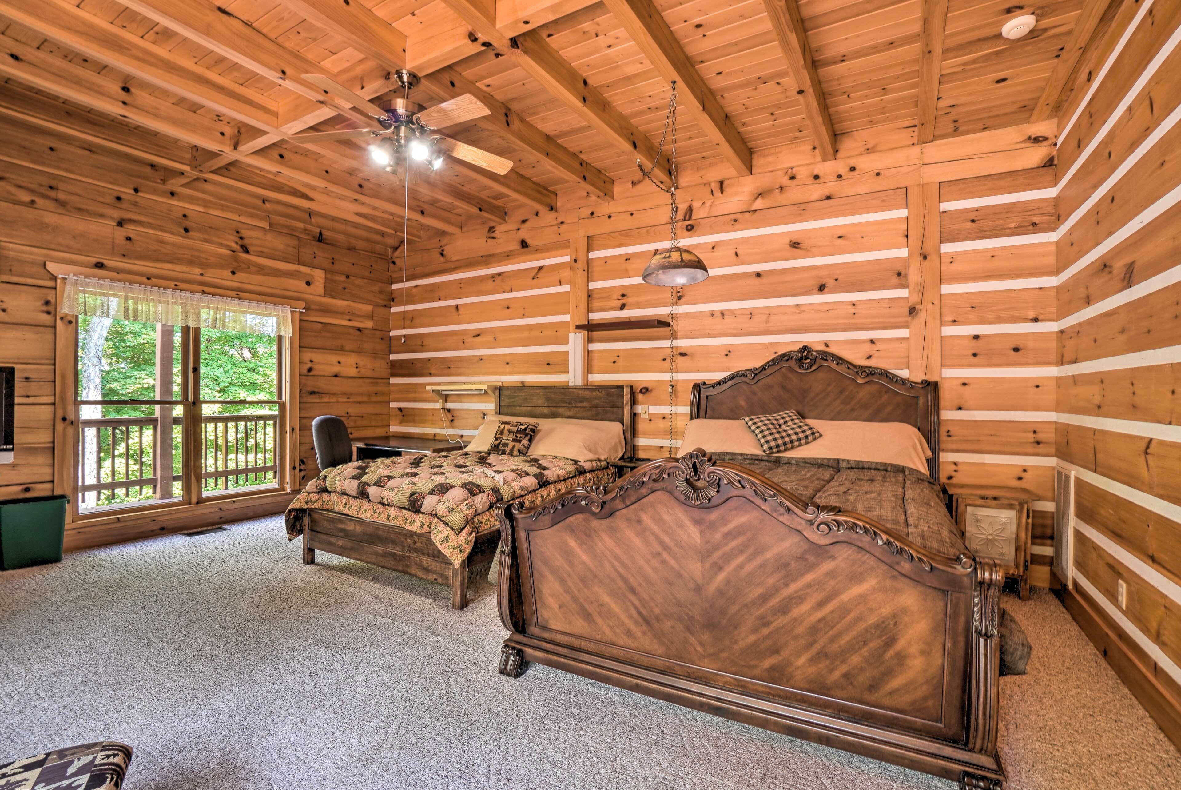 A log cabin bedroom with two wooden beds, a ceiling fan, and a large window with a view of greenery.