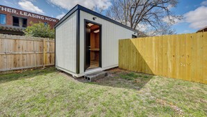 A small, single-story wooden structure with a door open, set against a backdrop of a wooden fence and a building with a 'leasing now' sign. - Home: Grassy Yard, Walk to Bentonville Square (Bentonville)