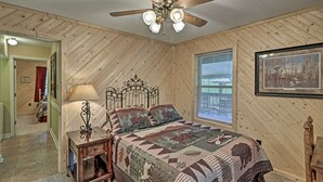 A bedroom with a wooden sloped ceiling, a bed with a quilt, a bedside table with a lamp, and a window with blinds.