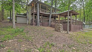 A two-story cabin with a balcony and a covered porch, surrounded by trees and a stone wall.