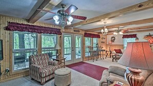A cozy cabin interior with a fireplace, wooden walls, a ceiling fan, and a large window overlooking a forest.