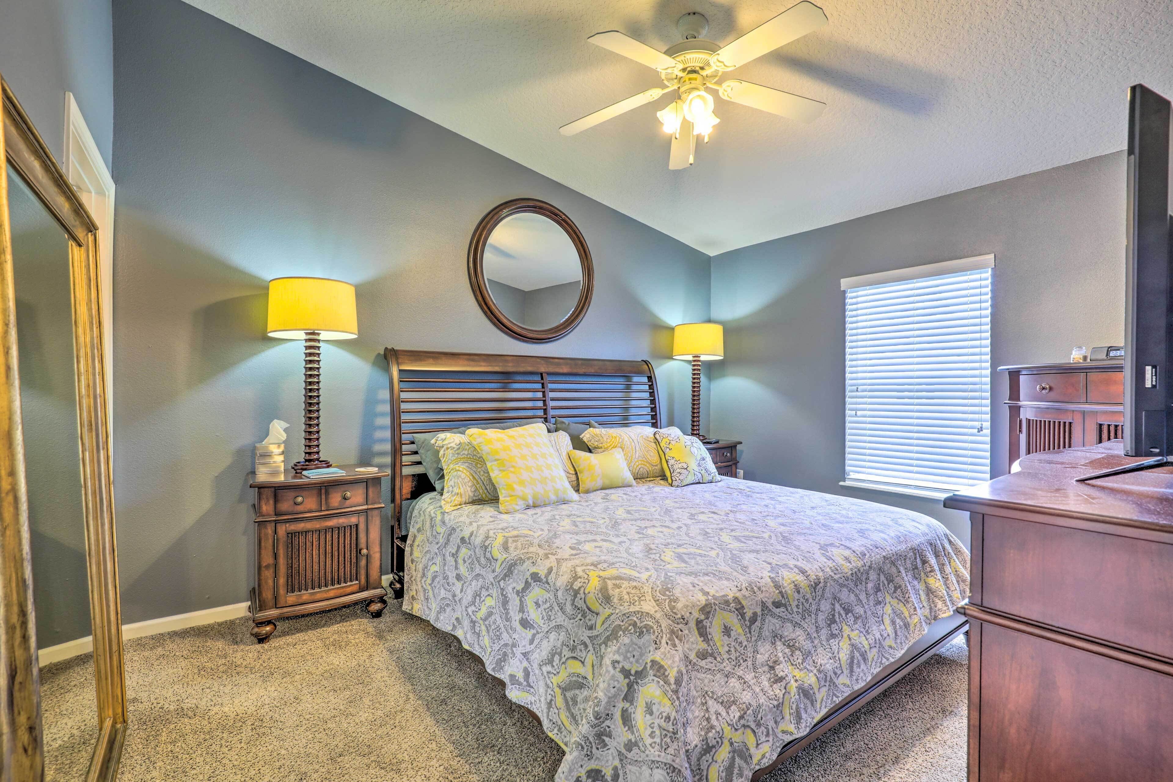 A bedroom with a bed, two bedside tables with lamps, a ceiling fan, a mirror, and a window with blinds.