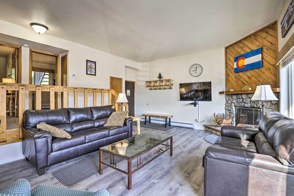 A living room with a brown leather sofa, a glass coffee table, and a stone fireplace. - Silverthorne Condo w/ Views ~ 11 Mi to Ski Slopes (Silverthorne)