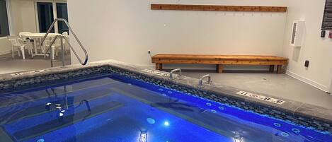 A swimming pool with a wooden bench and a table with chairs, a staircase, and a wall-mounted light fixture.