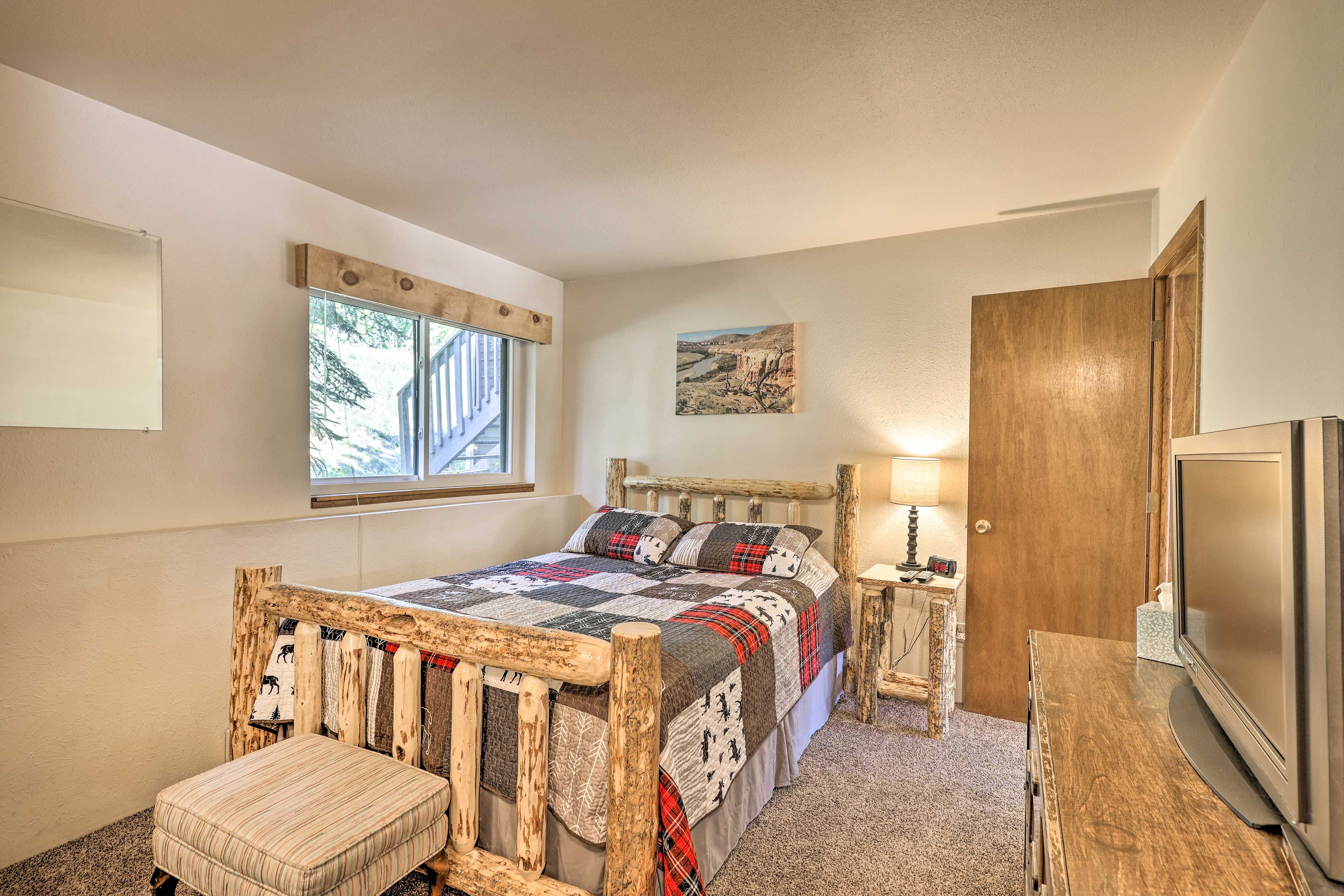 A bedroom with a wooden bed, a bedside table with a lamp, a TV, and a window with a view of trees.
