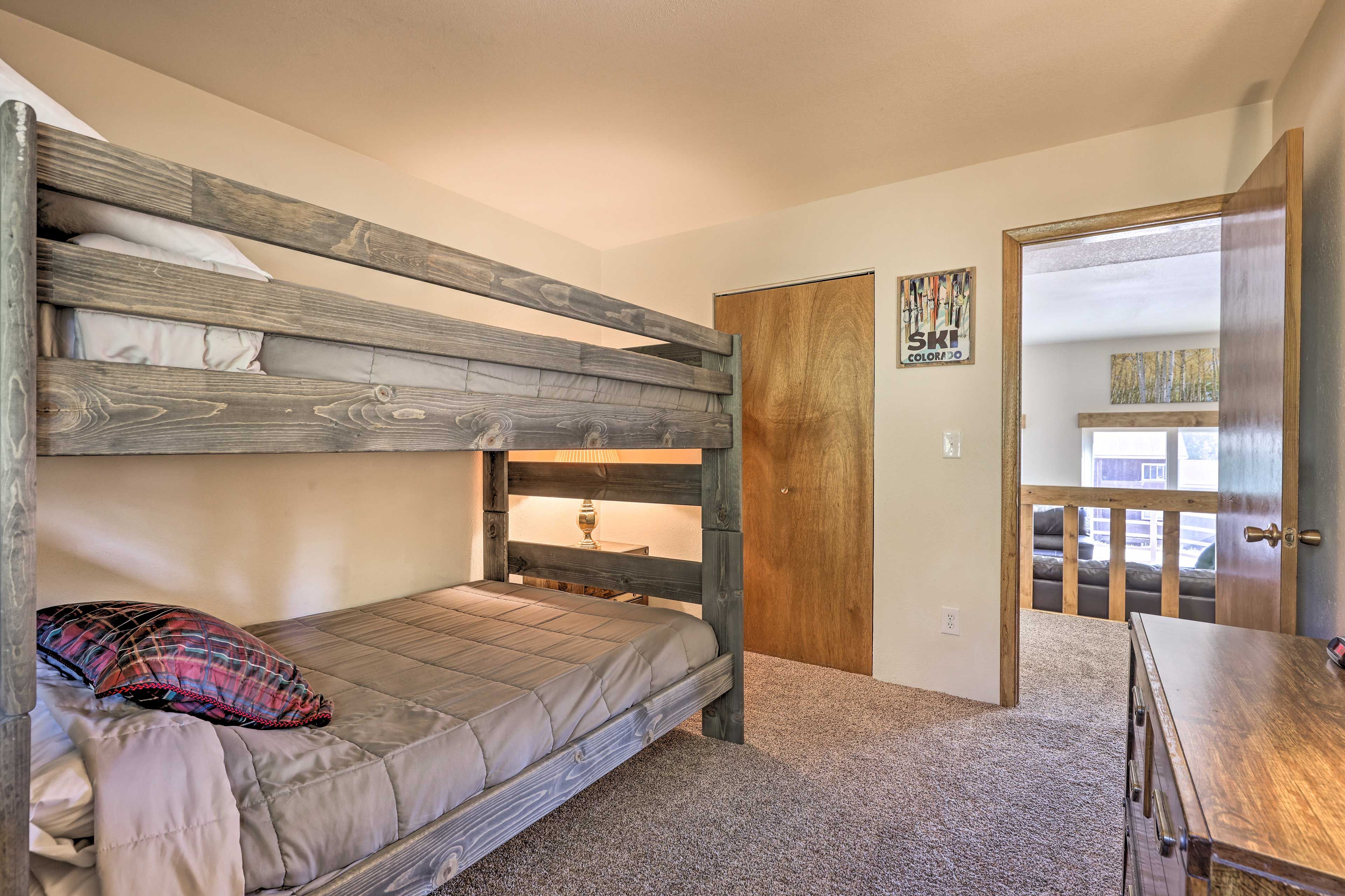 Una cama litera con cabecera de madera, una cama individual con almohada a cuadros, un aparador de madera y una puerta que da a un balcón.