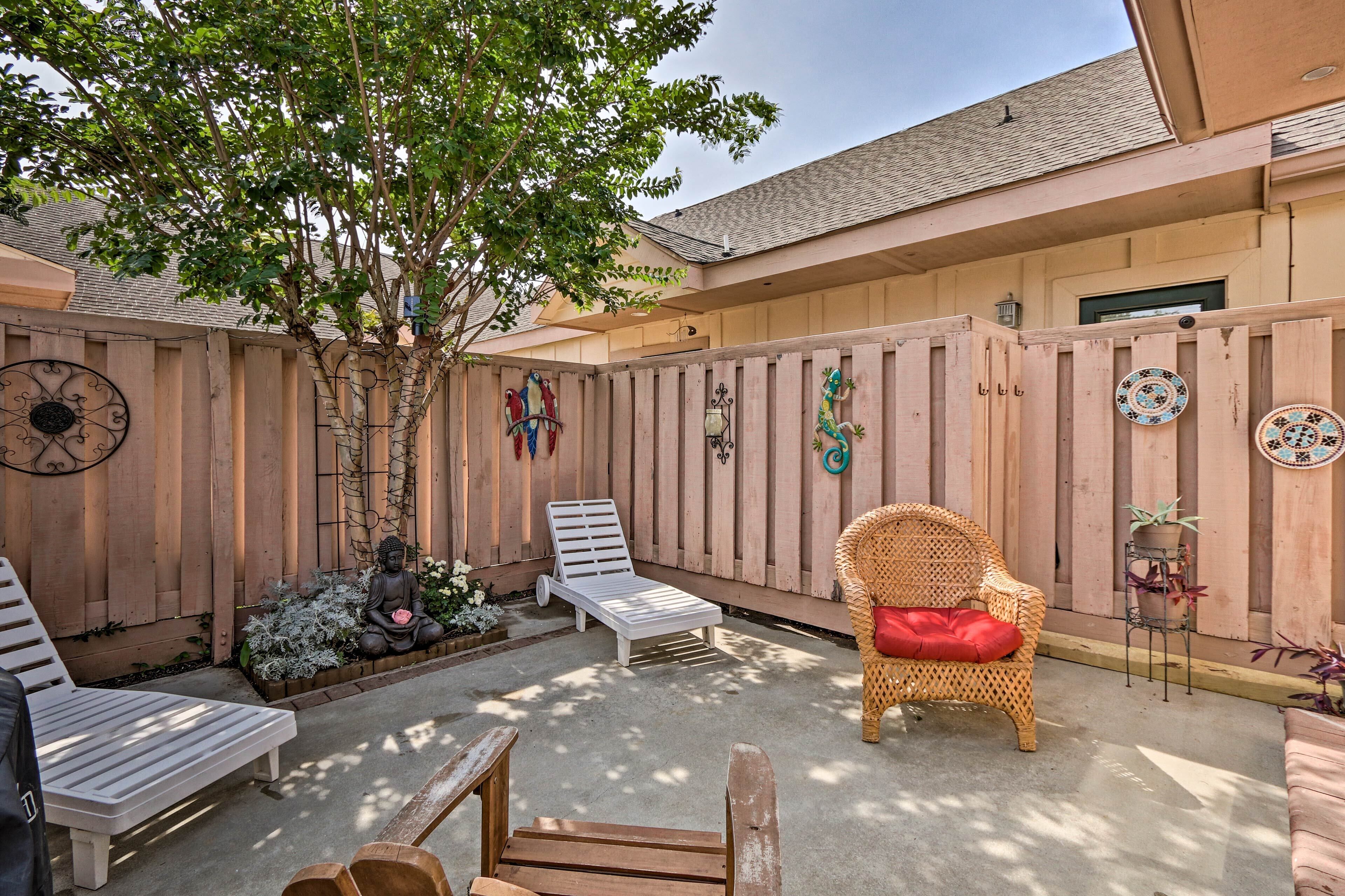 A backyard with a wooden fence, a statue, a white lounge chair, a wicker chair with a red cushion, and a small potted plant.