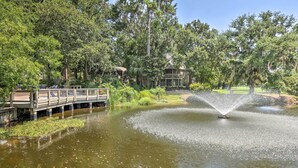 A serene pond with a fountain, surrounded by lush greenery and a wooden deck with lounge chairs.