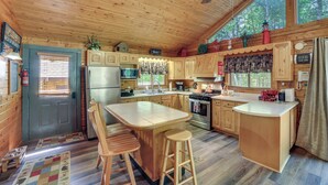 Cocina de cabaña de madera con una isla central, taburetes de bar, armarios de madera, refrigerador, estufa, microondas y un ventanal grande.