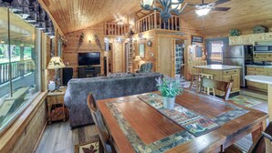 Interior de una cabaña de madera con comedor, sofá, televisión, cocina y un balcón con vistas.