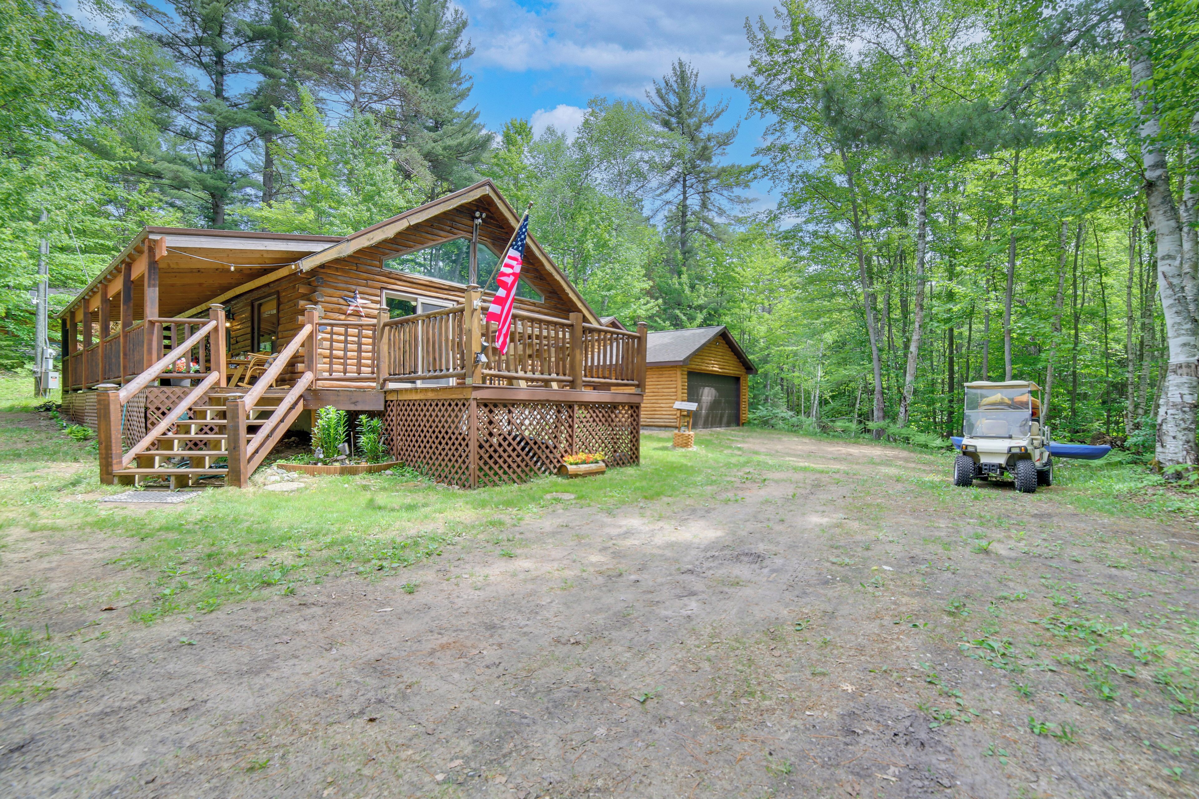 A wooden cabin with a deck, a flag, and a vehicle in a forested area.