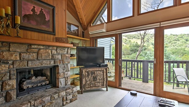 A stone fireplace with a television on a wooden stand, a wooden deck with a chair, and a view of a forested area. - Year-round Fun in Beech Mountain! Condo w/ Balcony (Banner Elk)