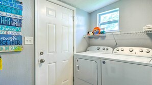 A laundry room with a front-loading washer and dryer, a window above the dryer, and a shelf with laundry supplies. - Charming Coastal Getaway, Near Beach & Pier! (Gulfport)