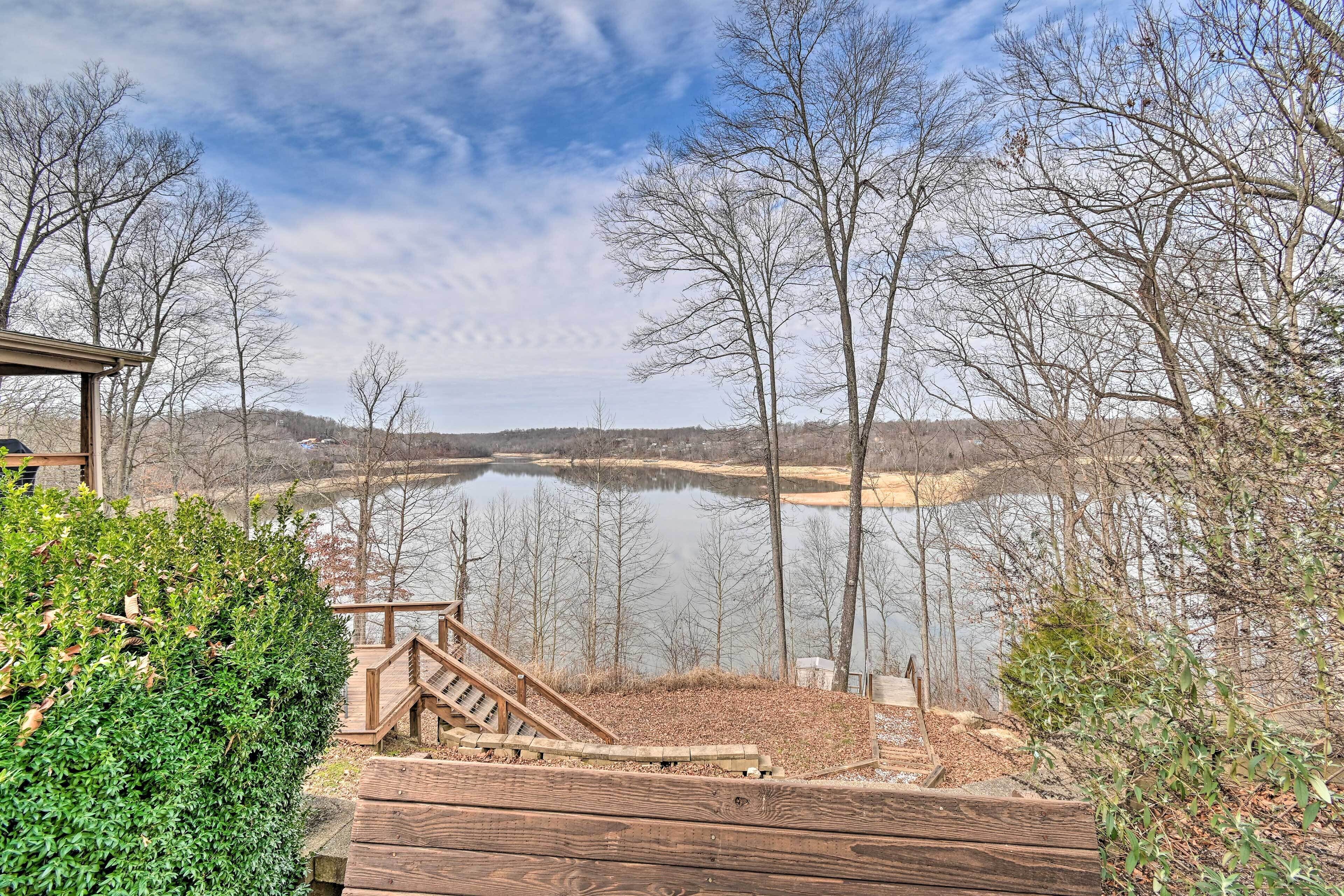 A wooden deck with a staircase leading to a viewing area overlooking a lake, surrounded by trees and a clear sky.