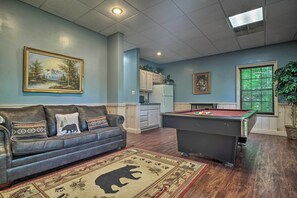 Uma sala de estar com mesa de bilhar, um sofá com almofadas decorativas, um quadro na parede e uma área de cozinha ao fundo.