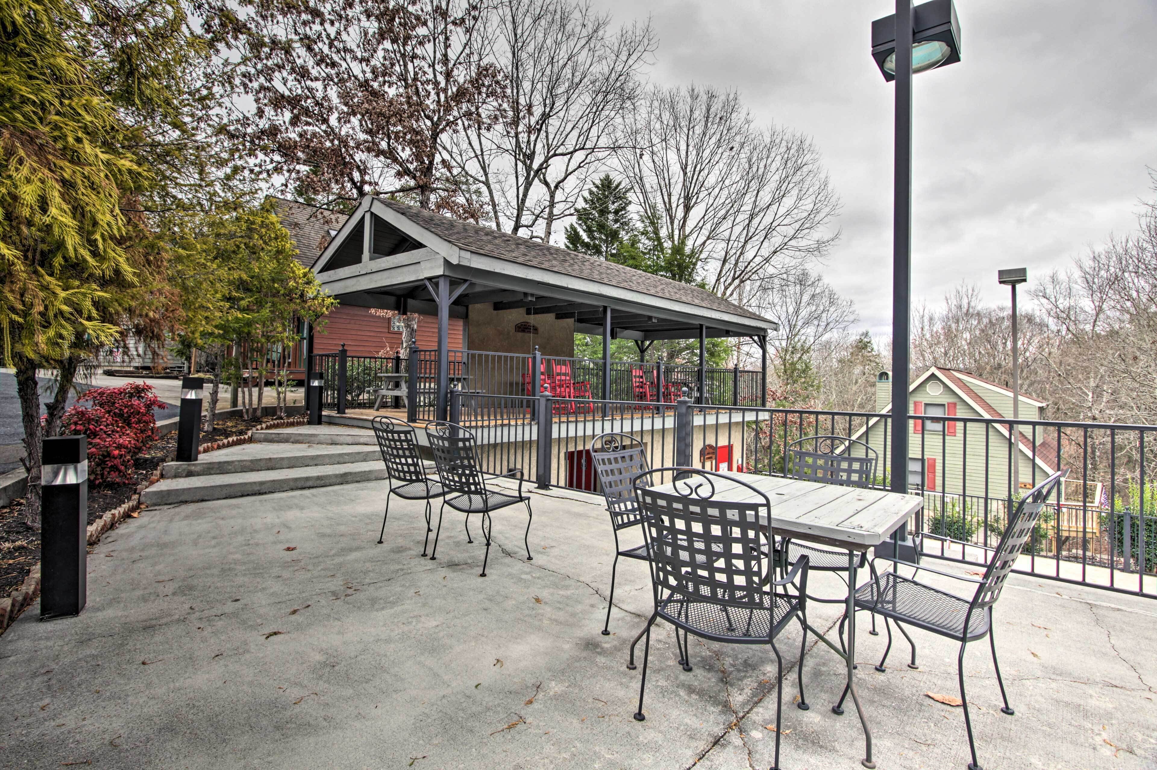 A covered outdoor seating area with metal chairs and a table, surrounded by a fence and trees.