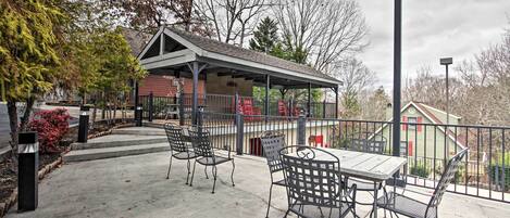 A covered outdoor seating area with metal chairs and a table, surrounded by a fence and trees.