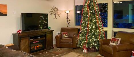 A living room with a fireplace, a television, a Christmas tree, and two armchairs.