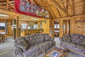 A cozy cabin interior with a wooden ceiling, a large quilt hanging, a floral sofa, and a kitchen area. - Rustic River View Cabin w/ Fire Pit, Games & Grill (Falmouth)