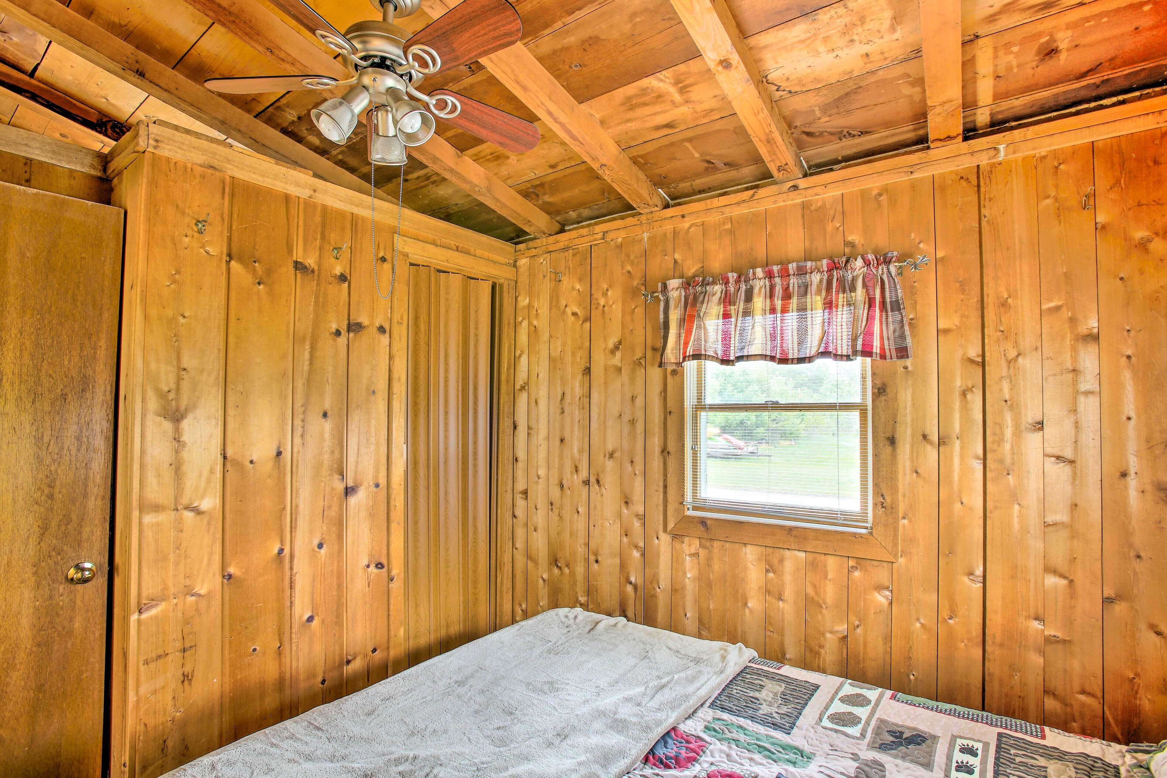 A wooden cabin room with a bed, a ceiling fan, and a window with a plaid curtain.