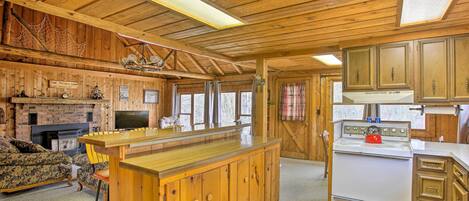A cabin interior with a wooden dining area, a fireplace, a kitchen with a stove, and a large window.