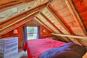 A bedroom with a sloped ceiling, wooden walls, a bed with a red and blue blanket, a dresser, and a window with curtains. - Rustic River View Cabin w/ Fire Pit, Games & Grill (Falmouth)