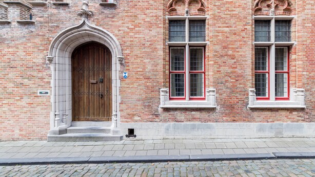 A historic building with a large wooden door, two windows with red frames, and intricate brickwork. - Room of Puebla (Brugge)