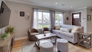 A living room with a sofa, two armchairs, a coffee table, and a small side table. There are curtains on the windows, a flat-screen TV mounted on the wall, and a wooden cabinet with plants.