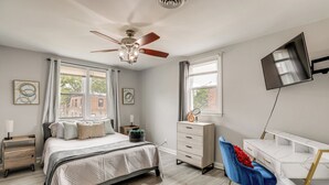 Un dormitorio con cama, cómoda, escritorio, silla, ventilador de techo y televisión.