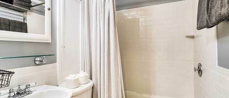 Un baño con lavamanos, inodoro y bañera blancos. Hay una cortina de ducha, un perchero y un espejo sobre el lavamanos.