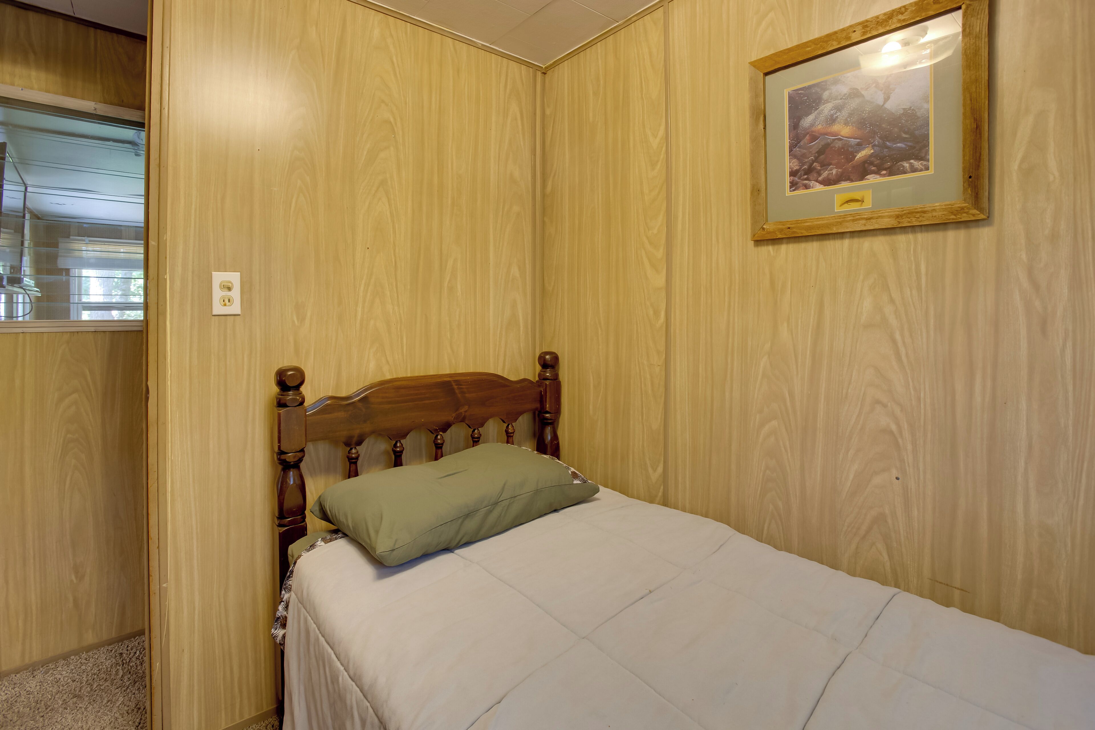 Un lit simple avec une tête de lit en bois et un oreiller vert, dans une pièce aux murs recouverts de lambris en bois et ornée d’un tableau encadré.