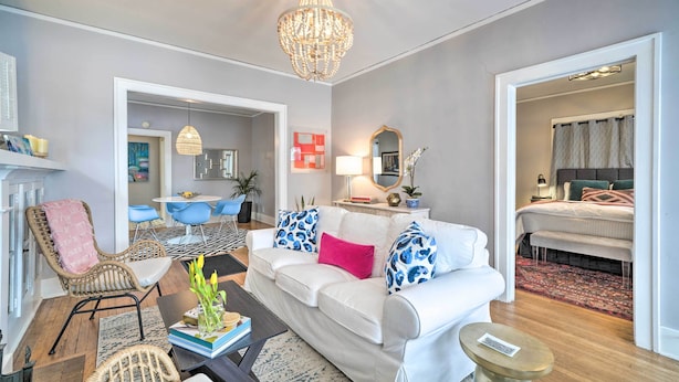 Uma sala de estar moderna com um sofá branco, almofadas estampadas em azul e rosa, uma cadeira de vime, uma mesa de centro e um lustre.
