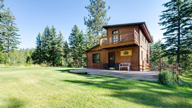 A two-story wooden cabin with a balcony, surrounded by a grassy yard and tall trees. - Conconully Cabin on 42 Private Acres Near Hiking! (Conconully)
