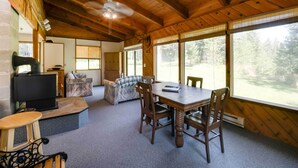 A cozy cabin interior with a dining table, chairs, a fireplace, and large windows offering a view of the forest. - Conconully Cabin on 42 Private Acres Near Hiking! (Conconully)