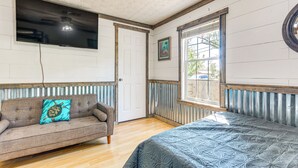 A room with a wooden floor, a sofa with a turtle-patterned pillow, a bed with a patterned bedspread, a mounted TV, a door, and a window with a view of the outside.