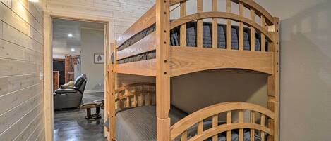Uma cama beliche com estrutura de madeira e colchão acolchoado, uma poltrona de couro e uma pequena mesa em um quarto com paredes de madeira e piso encerado.