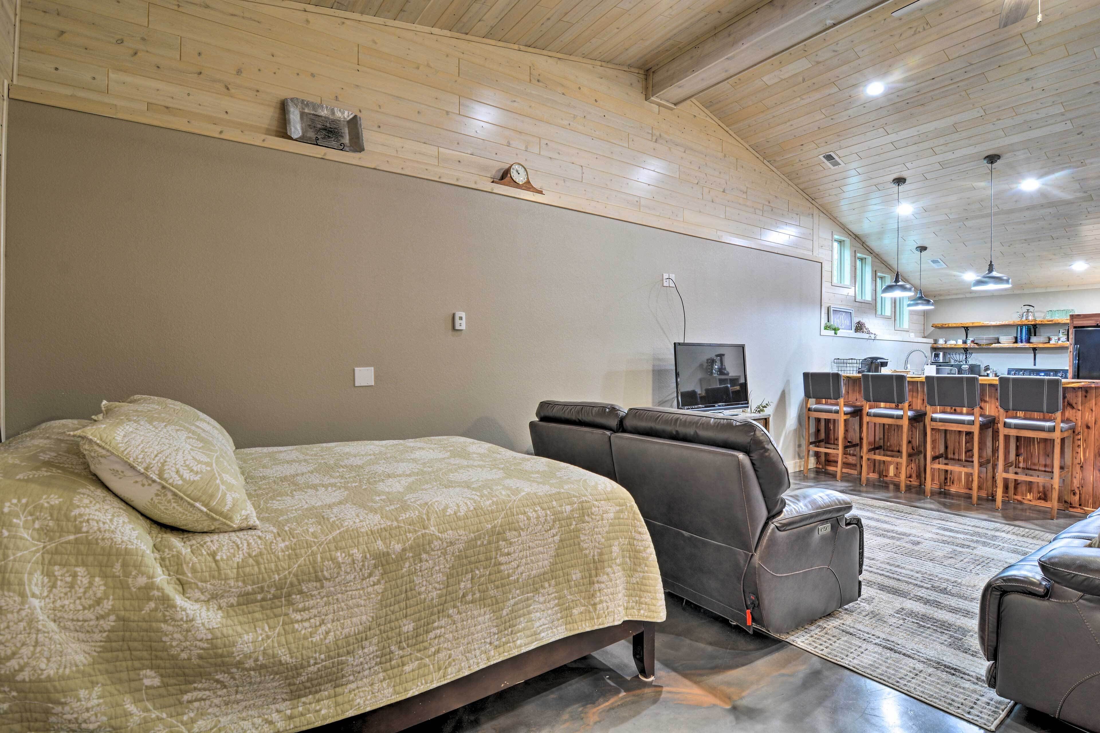 Una habitación con cama, una butaca reclinable, televisor y una zona de cocina con taburetes de bar.