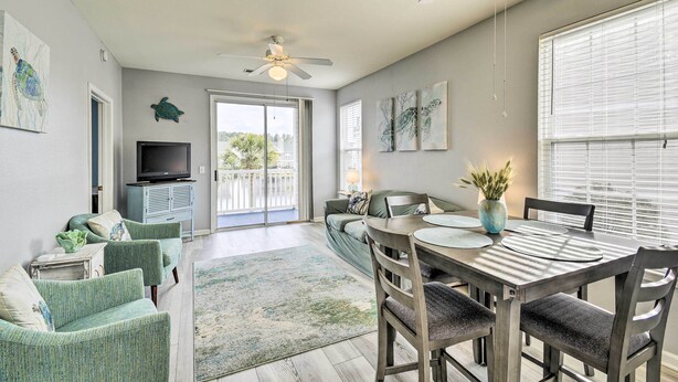 A spacious living room with a large window, a ceiling fan, a flat-screen TV, and a dining area with a table and chairs. - Sunset Beach Resort Condo in Golf Community! (Sunset Beach)