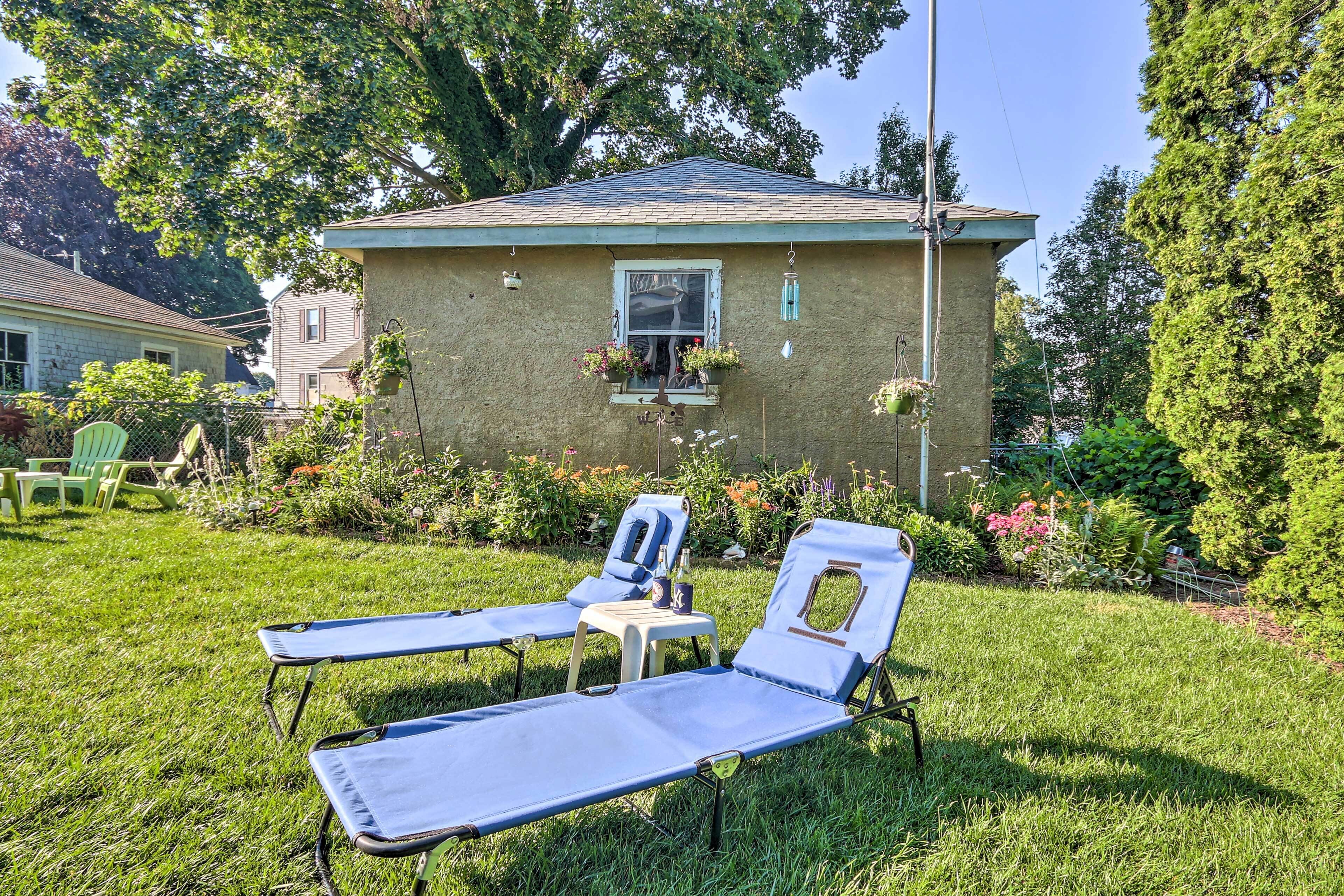 A house with a small garden, two lounge chairs, and a window with hanging plants.