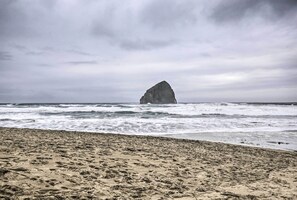 A beach with a large rock formation in the ocean, waves crashing onto the shore, and a cloudy sky. - Walk to Beach: Pacific City Home w/ Bay Views! (Pacific City)