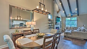 A spacious dining area with a large wooden table, chairs, and a chandelier. There is a kitchen in the background with a stone wall and a fireplace. - Spacious Family Home w/ Fire Pits, Large Yard (Goshen)