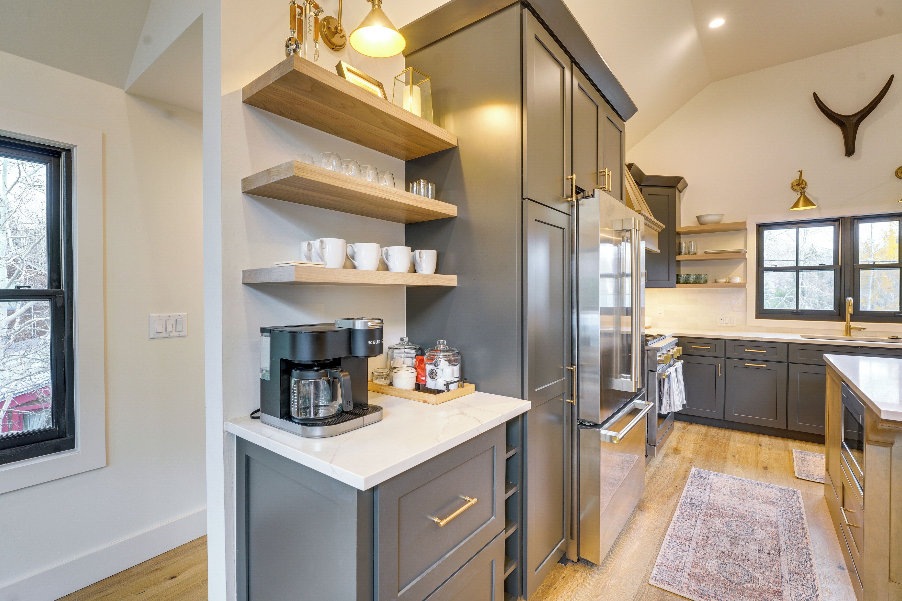 A modern kitchen with grey cabinets, a white countertop, and built-in appliances. There are wooden shelves above the counter and a large window on the left.