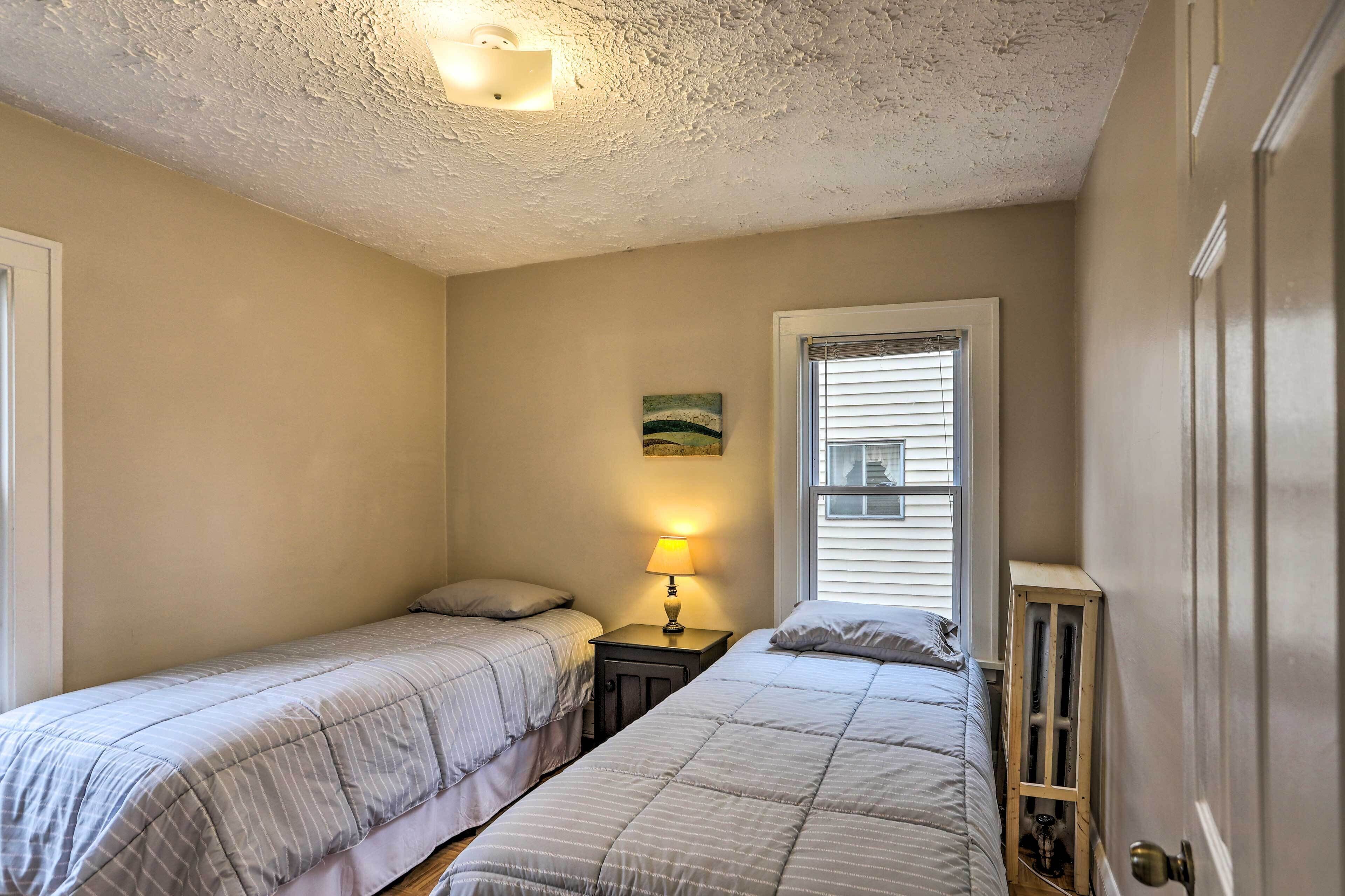 A bedroom with two single beds, a nightstand with a lamp, a window with blinds, and a door with a radiator.