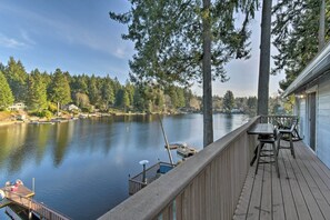 Een houten terras met uitzicht op een rustig meer, afgemeerde boten en een rij huizen op de achtergrond.