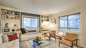 A cozy living room with a white sofa, a brown leather chair, a glass coffee table, and a bookshelf filled with books and decorative items. - Stunning Evergreen Chalet w/ Private Hot Tub! (Evergreen)